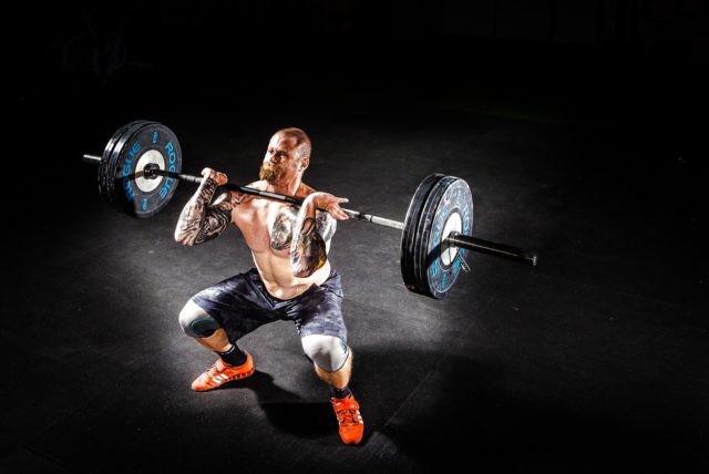 This photo shows a shirtless, heavily tattooed man in a deep squat. A barbell with heavy-looking weights is resting on his chest. He appears to have a spotlight on him, which illuminates only a small sliver of the floor. The rest of the background is lost in shadows.