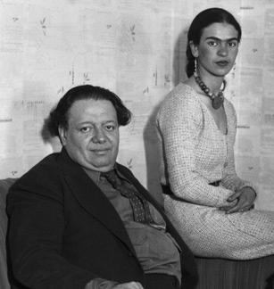 This undated black-and-white photo shows Diego Rivera and Frida Kahlo together. Rivera is seated on the left. He is dressed in a black jacket, shirt, and tie. His hair has grown past his ears. He's a portly fellow, of perhaps 300 pounds. Kahlo is perched on a higher surface (either the arm of a couch or a high stool) to his left. She's wearing a white dress, earrings, and a chunky necklace.