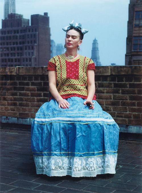 This photo of Frida Kahlo was taken by her lover, Nickolas Muray, in 1939. Kahlo sits on a rooftop in New York City. A brick wall is visible behind her, and beyond that is the New York City skyline. Kahlo is presumably sitting on a chair, although the chair is obscured by her long blue-and-white skirt. She is wearing a patterned red-and-yellow tunic in the Tehuana style, although she's missing the long, rectangular shawl that often completes the look. Her hair is done in an elaborate style with two large, blue bows that match her skirt. She wears chunky earrings and a large necklace. Her face is turned slightly to her right, and she holds a cigarette in her left hand.