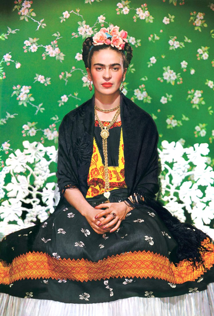 This photo of Kahlo was taken by her lover, Nickolas Muray, in 1939. Frida sits on a white bench with a patterned, floral back. The wallpaper behind her is green, and has a floral print that goes remarkably well with the arrangement of pink flowers in Kahlo's hair. Kahlo is wearing a black shawl over a patterned yellow, orange, and black shirt, and a long skirt with flowers and a white ruffle at the bottom. She's wearing a long gold necklace, earrings, and a large rind.