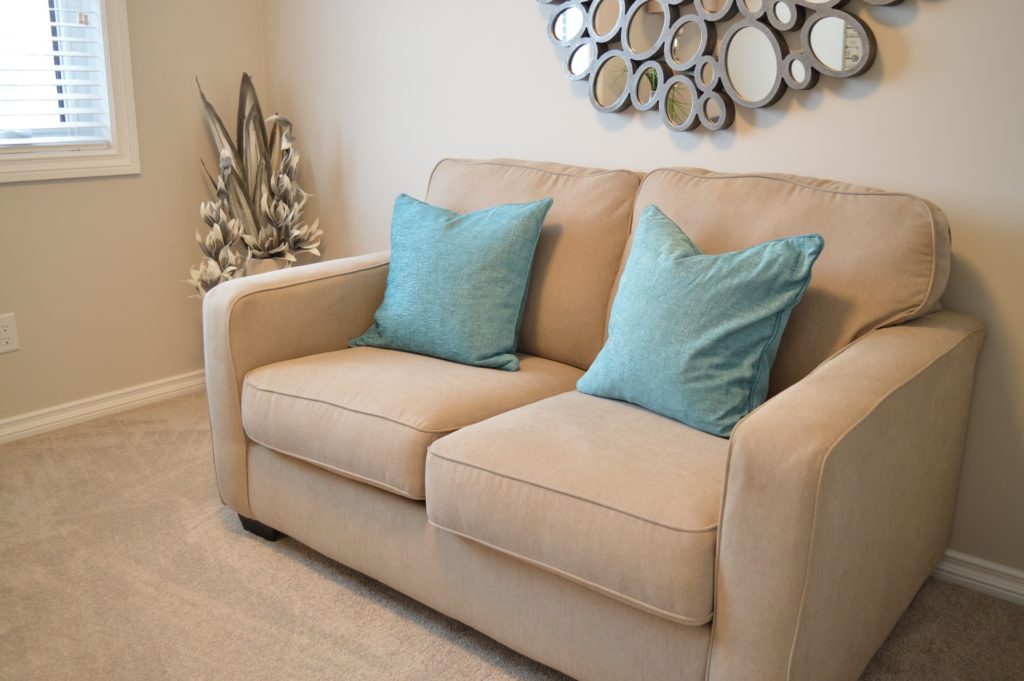 A photo of a cream-colored, cushy loveseat in an uncluttered, cream-colored living room. The walls and carpet are beige, and a weird artsy mirror is hanging on the wall behind the loveseat. Two blue cushions on the loveseat provide the only notes of color.