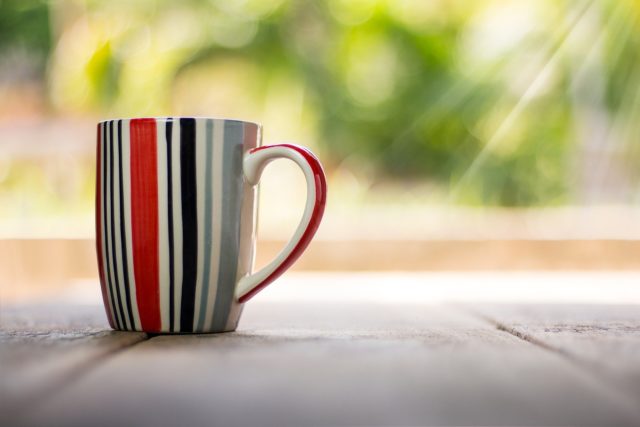 This photo shows a striped coffee mug on top of a wooden table. The background is blurred out, but there is greenery out there, and carefully angled rays of sun. I suspect this photo was meant to illustrate, "Good Morning!"