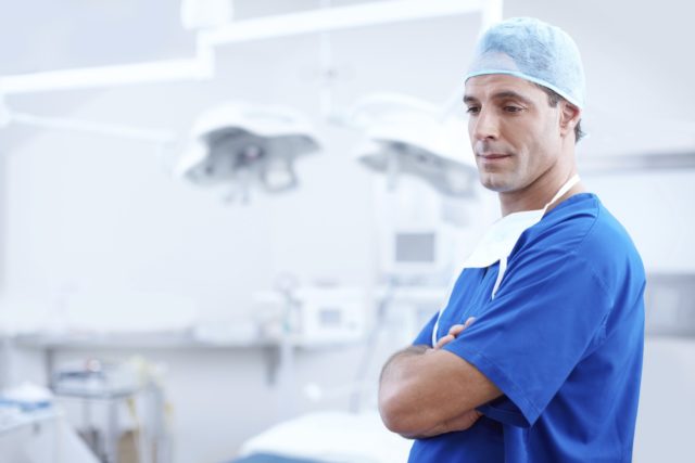 A surgeon (or more likely, an actor pretending to be a surgeon) is standing in blue scrubs, with arms crossed, and a slight smile on his face. A white, sterile-looking operation room is just visible behind him, although the background is blurred to allowed him to stand out in better focus.