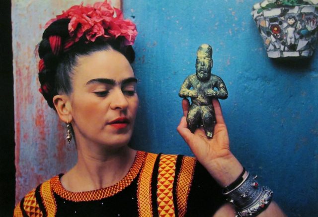 This photo of Frida Kahlo shows her posing in front of a blue wall, holding a carved stone statue in her left hand. The photo is a close-up, and shows her from the upper chest to the top of her head. She is wearing a black, yellow, and orange patterned shirt, thick metal bracelets, and long, dangling earrings. Her hair is twisted and braided into her signature style, with pink flowers woven in. She is looking slightly to her left, toward the statue.