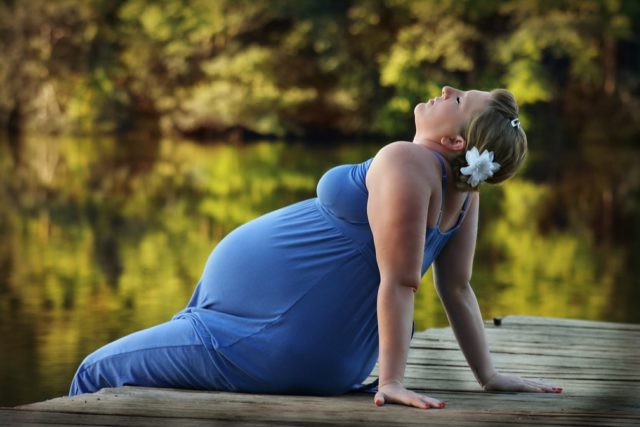 A pregnant woman sits on a rustic wooden pier. Her hands are both on the pier behind her, and she arches her back as she leans backwards. She has a flower in her hair, and a rather peaceful expression on her face, so I take it this feels good. The trees and river are in soft focus in the background.