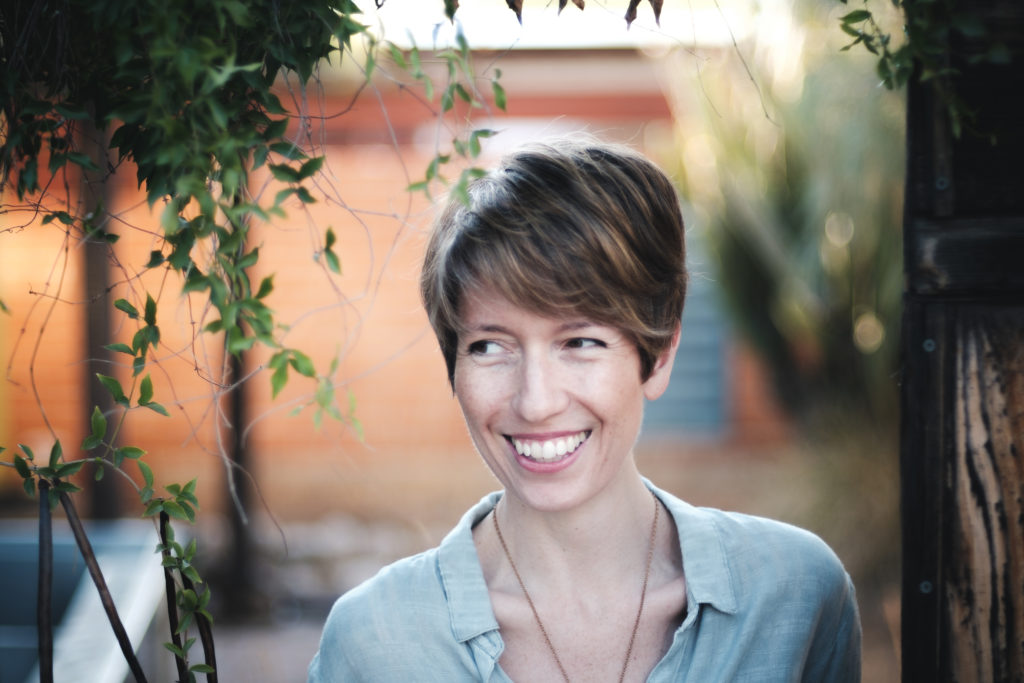 A photo of Sarah Ramey, author of The Lady’s Handbook for Her Mysterious Illness. It was taken outside, and some trees and greenery are mixed with urban features like a door and ledge in soft focus. Ramey is shown from the chest up. She's a white woman in her 30s with short brown hair and a wide smile. She's wearing a casual blue, collared shirt, and a gold chain for a necklace. (The pendant which presumably hangs from it is not in the shot.) Ramey is looking slightly off to her right, and grinning.