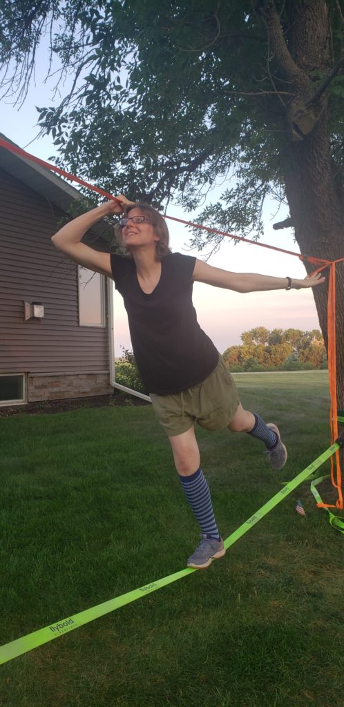 A photo of me at 19 weeks pregnant. I'm wearing a black v-neck tee shirt, army green shorts, striped compression socks, and tennis shoes. I'm walking across a slackline strung between two trees. My aunt's farmhouse and a row of trees are visible in the background.