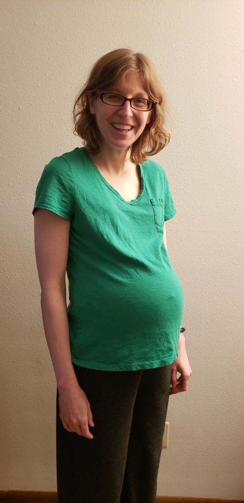 A photo of me at 38 weeks pregnant. I'm standing with my arms at my sides. I'm wearing a green v-neck tee shirt that shows off my baby bump.