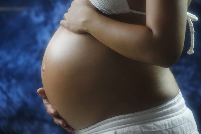 This photo is a close-up of a pregnant woman's bare belly. She is wearing white shorts and a white halter top, which are barely visible at the top and bottom of the photo. Her left hand rests on top of her belly, while her right hand cradles it from below.