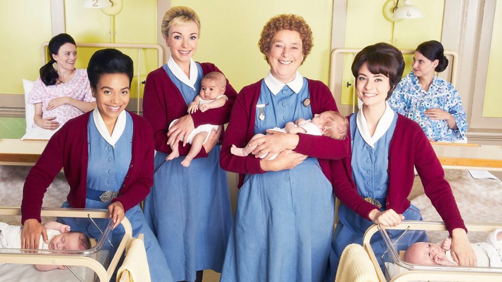 A promotional shot from the BBC's television series Call the Midwife. Four of the main characters, all of them midwives, stand front and center. They are wearing matching uniforms - blue dresses with red cardigans. The two midwives in the middle hold babies, while the two midwives on either side each place a hand on an infant in a bassinet. In the background, two pregnant women lying in hospital beds exchange smiles.