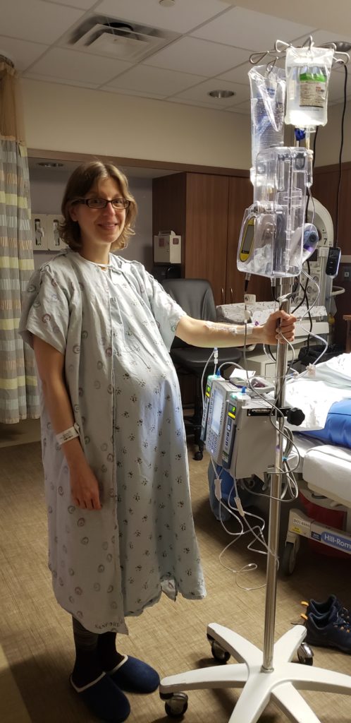 A photo of me in my hospital room during labor. I am wearing a hospital gown, and holding my IV pole in my right hand. When this photo was taken, I was already on Pitocin and a saline drip.
