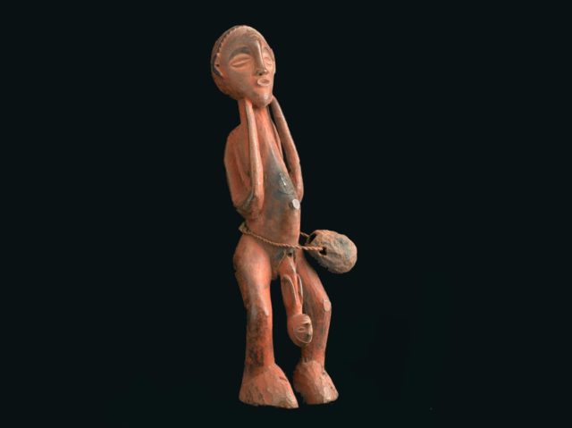 A photo of a carved wooden figure set against a black background. The figure is abstract, and shows a woman standing up, while a baby slides out headfirst from between her legs. The woman's elbows are at her sides, while her hands (really, the sticks where her hands should be), are under her chin. There is a rope slung around her waist with a gourd hanging from it.