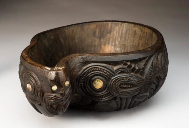This is a photo of a carved wooden placenta bowl. The shape is weird, something like a twisted teardrop. The carvings around the outer rim are abstract, with lots of loops and whirls. It is also inlaid with pieces of abalone shell.
