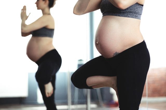 A pregnant woman stands in front of a mirror in a yoga studio. At least I assume it's a yoga studio, because she's doing tree pose. She's standing on one leg, with her other foot placed besides her knee. Her hands are held together. She's wearing a sports bra and yoga pants, and a tattoo is just visible in her pregnant midsection.