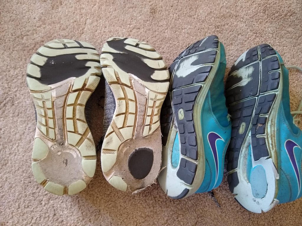 A photo of the bottoms of my two old pairs of tennis shoes. It's hard to say which pair is worse. Both are missing big chunks from the balls and heels.