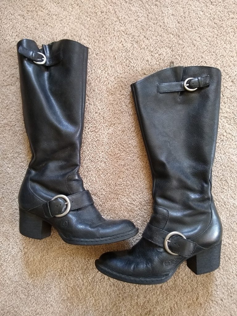 A pair of black, calf-high leather boots. Each boot has one silver buckle at the top, and another on the arch. The heels are about two inches high.