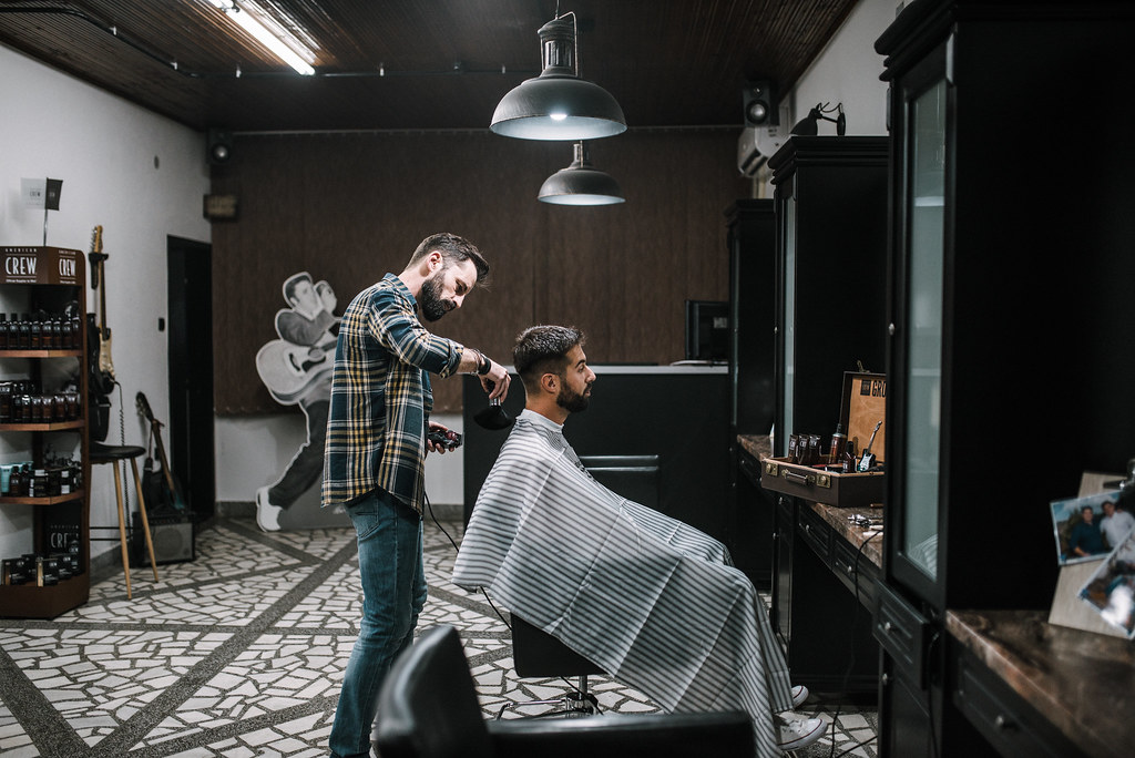 A male barber is cutting a second man's hair. They are standing in what appears to be a very hipster salon. There is a cardboard cutout of Elvis visible in the background, and the salon has a repurposed industrial aesthetic. The barber is quite well-groomed himself, and sports a plaid flannel shirt and skinny jeans.