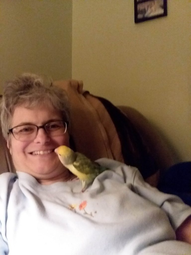 A photo of Maryanne laying back in a recliner. She's smiling, and a curious parakeet is poking around her mouth.