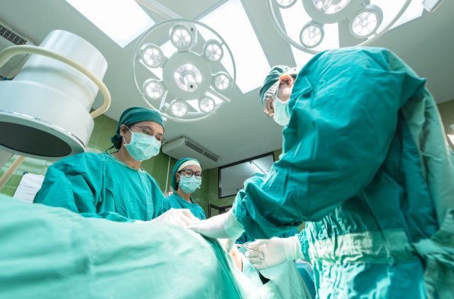 A surgeon and operating room staff stand poised over a patient's bed. The surgeon is standing to the right, holding a scalpel. The photo is taken from below the patient, so we get a good view of the ceiling and its many fluorescent lights, but we can't see the patient. Everyone is wearing green scrubs, and the medical people are wearing masks.